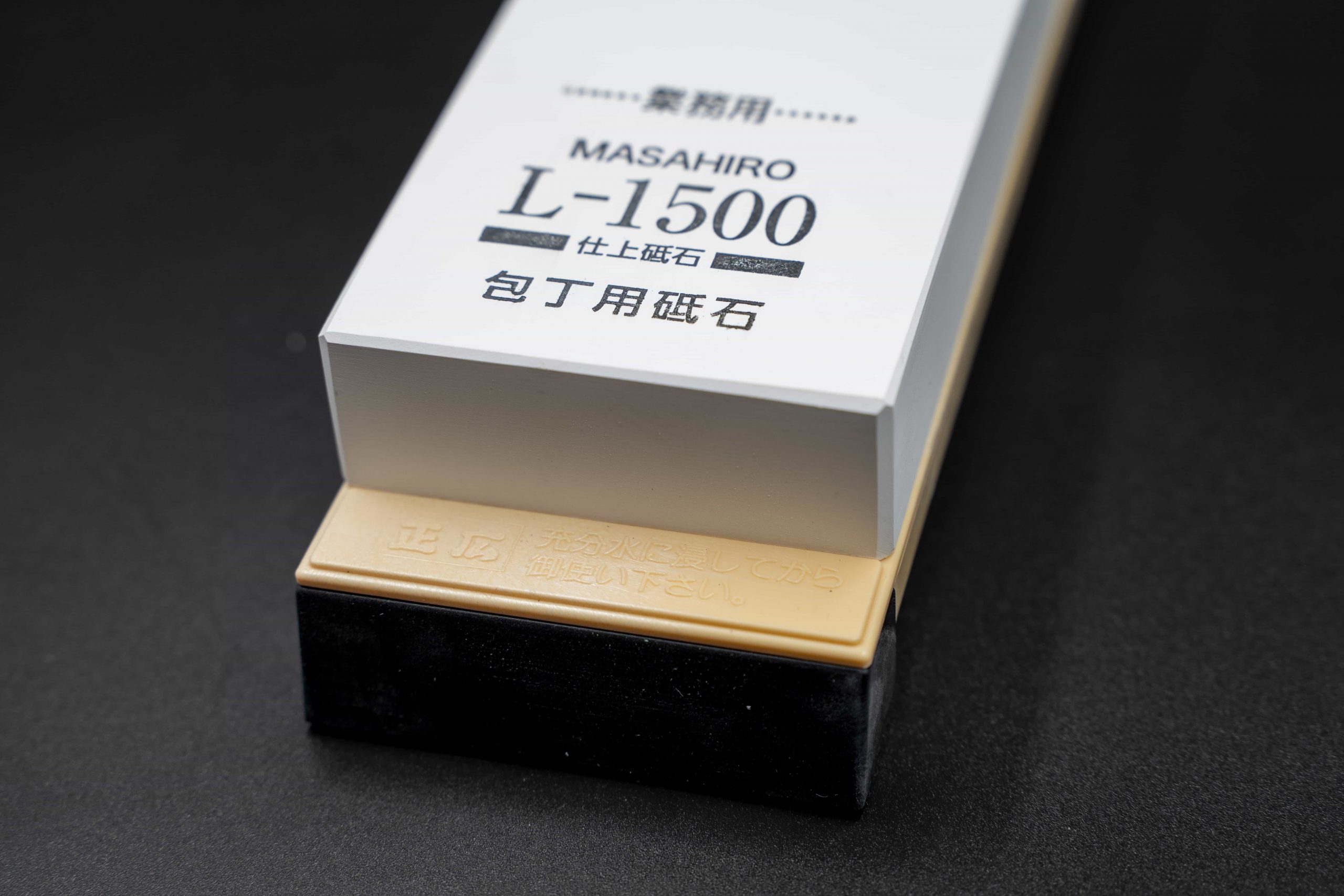 Ein rechteckiger Masahiro L-1500 Schärfstein mit japanischer Aufschrift liegt auf einer schwarzen Fläche neben einer beige-schwarzen JABA - Schneidebrett Drache Basis.