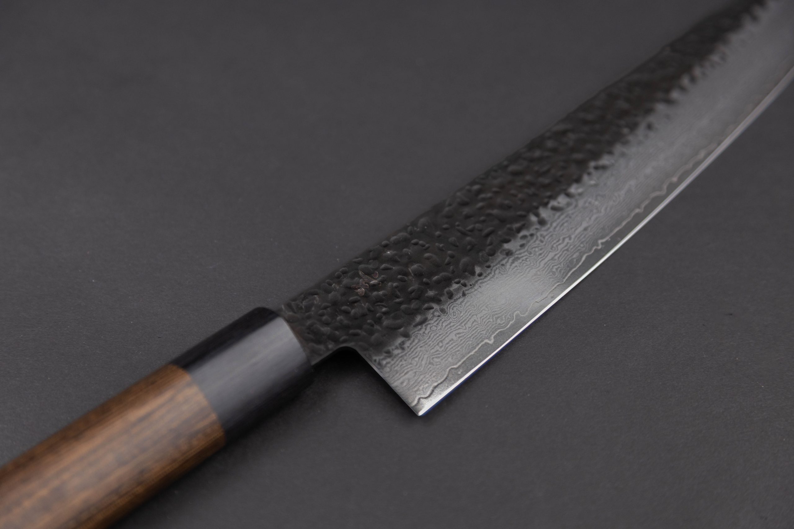 Eine Nahaufnahme des Gen - Gyuto 210mm Kochmessers mit dunklem Holzgriff und gehämmerter, gemusterter Stahlklinge vor einem schlichten, dunklen Hintergrund.