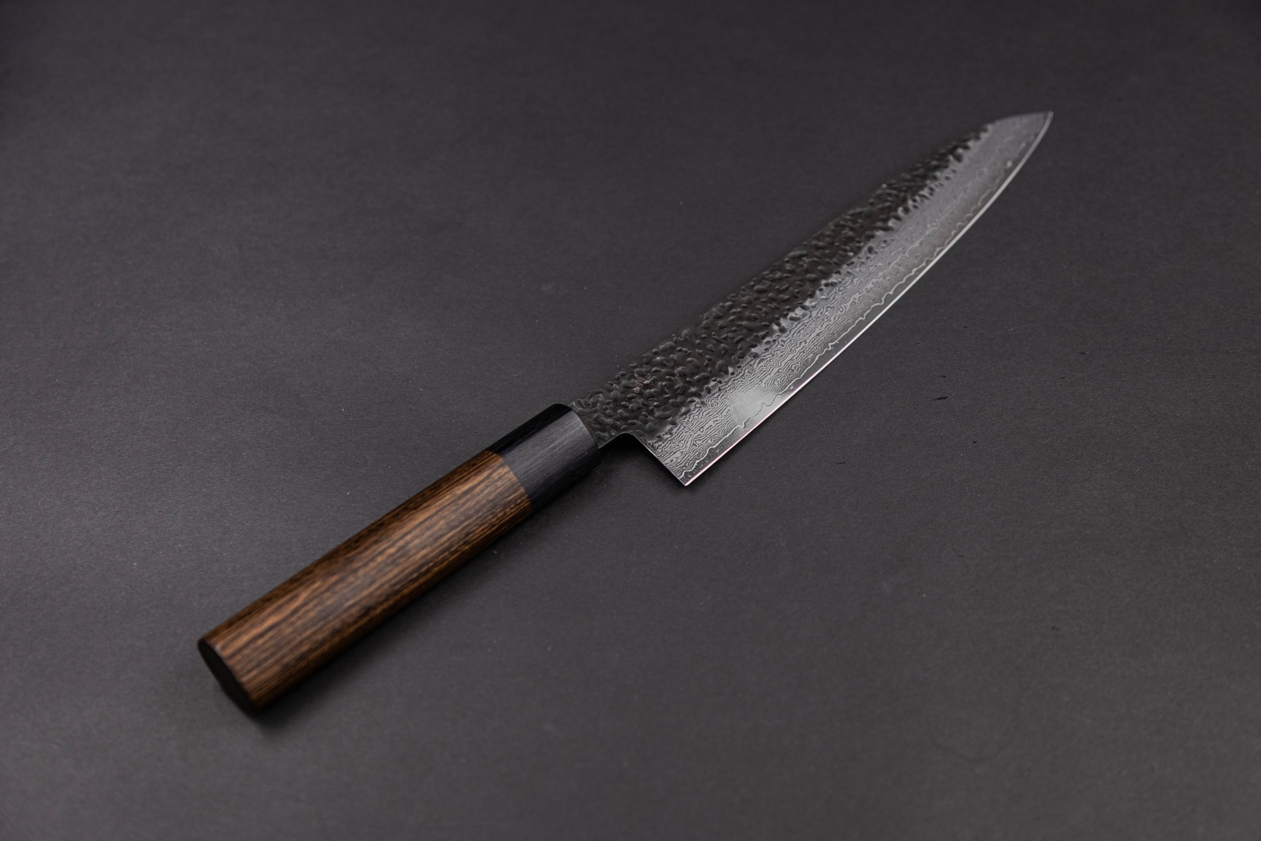 Das Gen - Gyuto 210mm, ein japanisches Kochmesser mit Holzgriff und gehämmerter Klinge, ruht auf einer dunklen Oberfläche - ideal für alle, die das Gen Gyuto kaufen wollen.