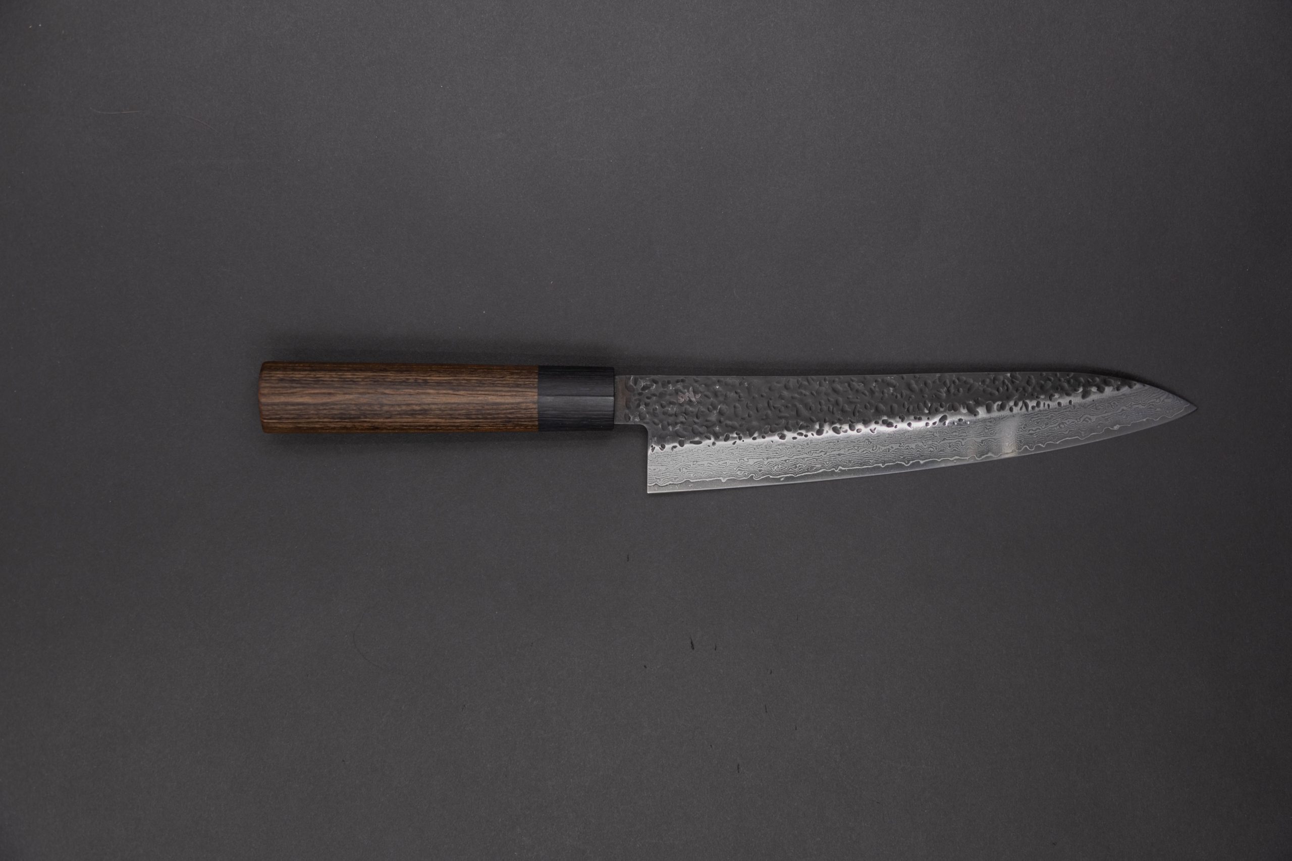 Das Gen - Gyuto 210mm ist ein Kochmesser im japanischen Stil mit einer Klinge aus gehämmertem Stahl und einem Griff aus dunklem Holz auf einem dunkelgrauen Hintergrund.
