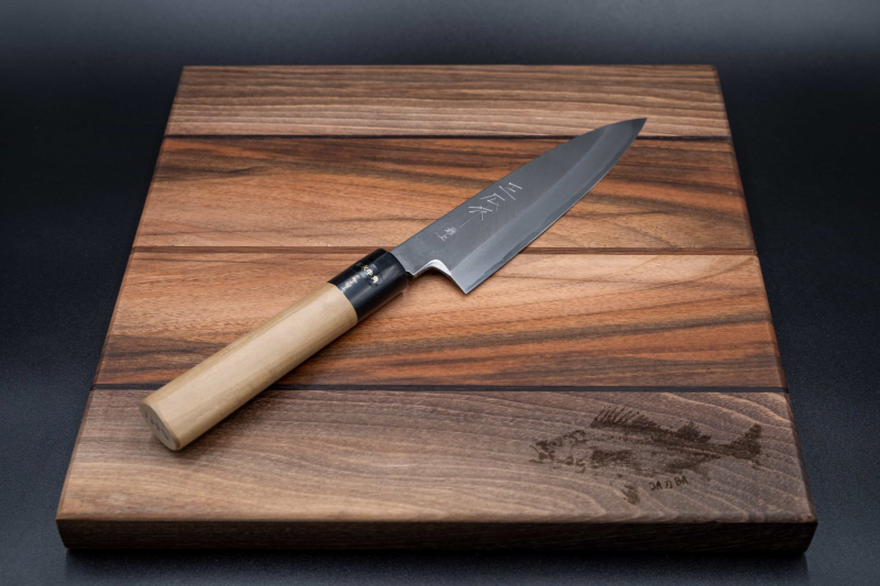 Ein 180-mm-Kochmesser von Masahiro - Funayuki mit Holzgriff liegt diagonal auf einem Holzschneidebrett mit einer Fischgravur in der Ecke, alles vor einem dunklen Hintergrund.