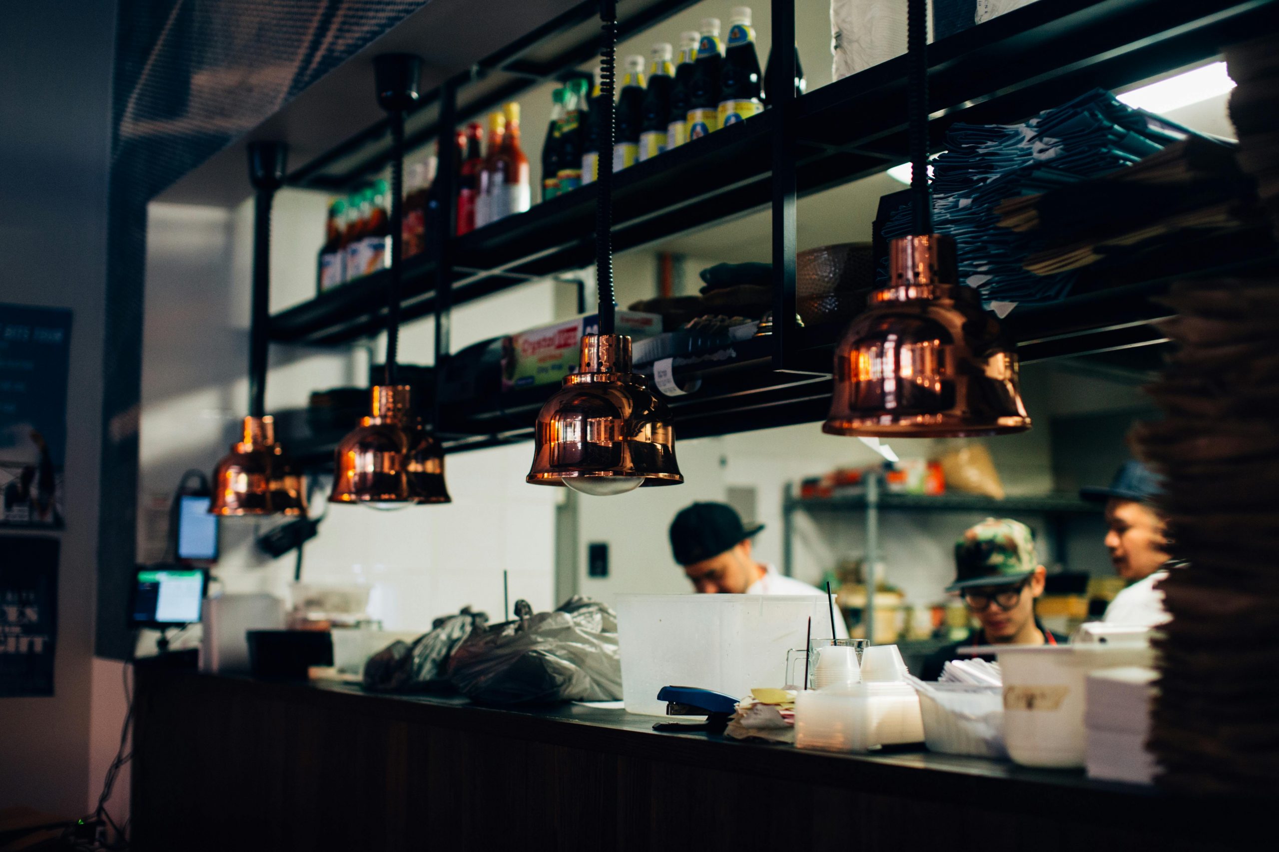 Eine Restaurantküche mit Köchen, die hinter einem Tresen arbeiten, beleuchtet von kupfernen Pendelleuchten. In den Regalen darüber stehen Flaschen, und im Vordergrund sind Teller und Küchenutensilien gestapelt.