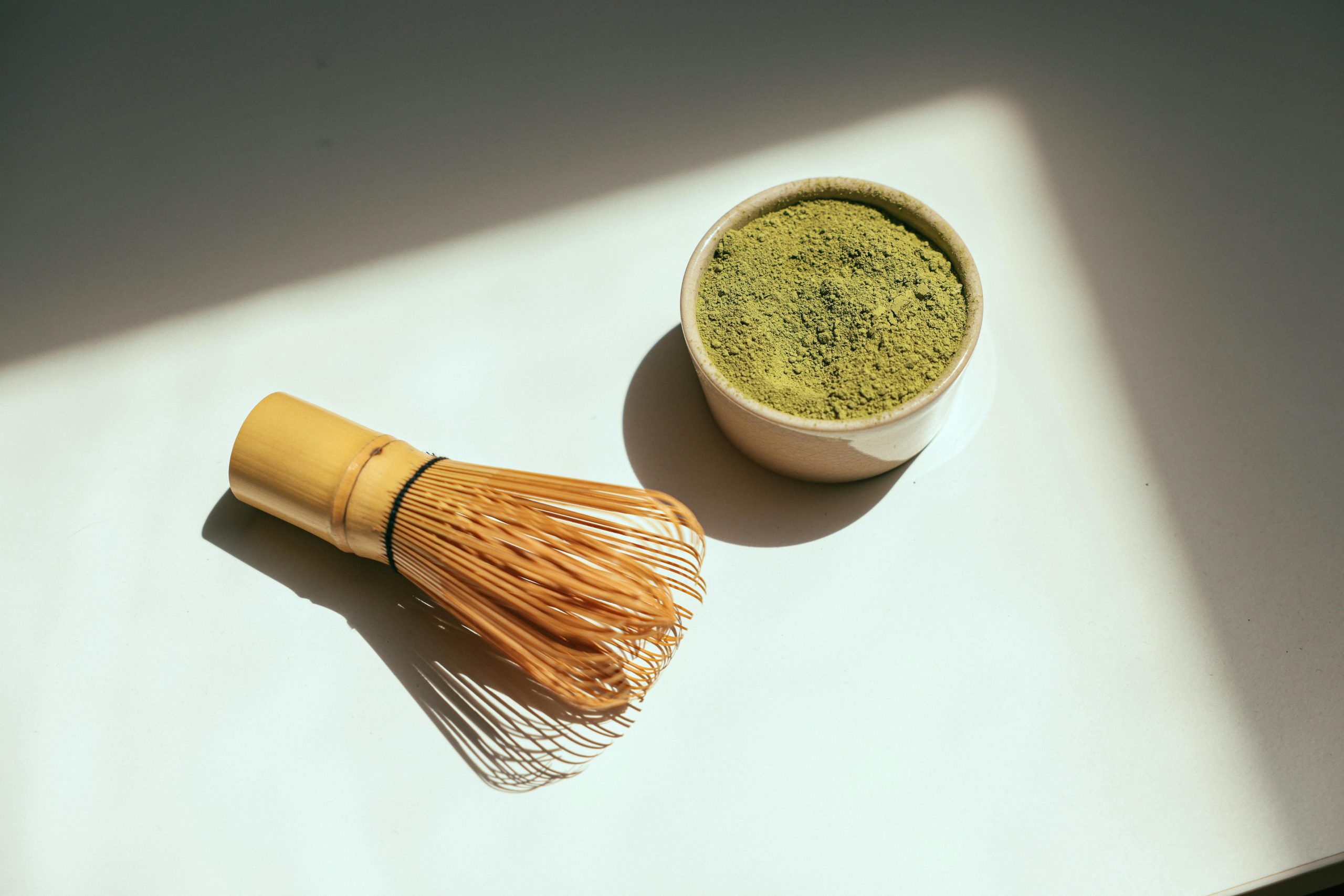 Ein Bambus-Matcha-Besen und eine kleine Schale mit grünem Matcha-Pulver stehen auf einer weißen Fläche, und das Sonnenlicht wirft Schatten auf die Szene.