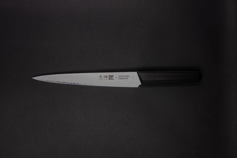 Ein Küchenmesser im japanischen Stil mit einem dunklen Griff und einer scharfen silbernen Klinge, die auf einem schlichten dunklen Hintergrund abgebildet ist. Auf der Klinge sind Text und Symbole eingraviert.