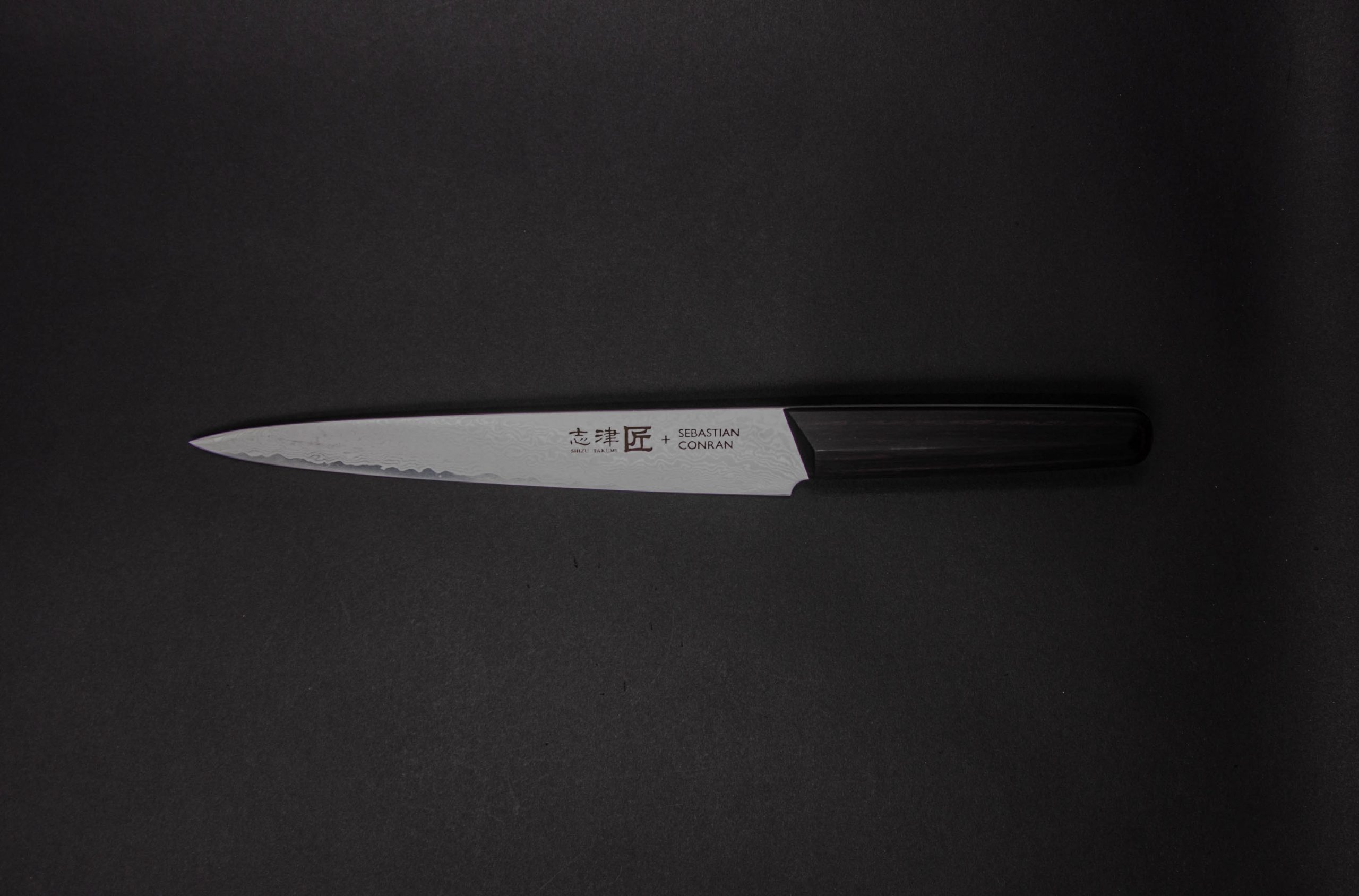 Ein Küchenmesser im japanischen Stil mit einem dunklen Griff und einer scharfen silbernen Klinge, die auf einem schlichten dunklen Hintergrund abgebildet ist. Auf der Klinge sind Text und Symbole eingraviert.