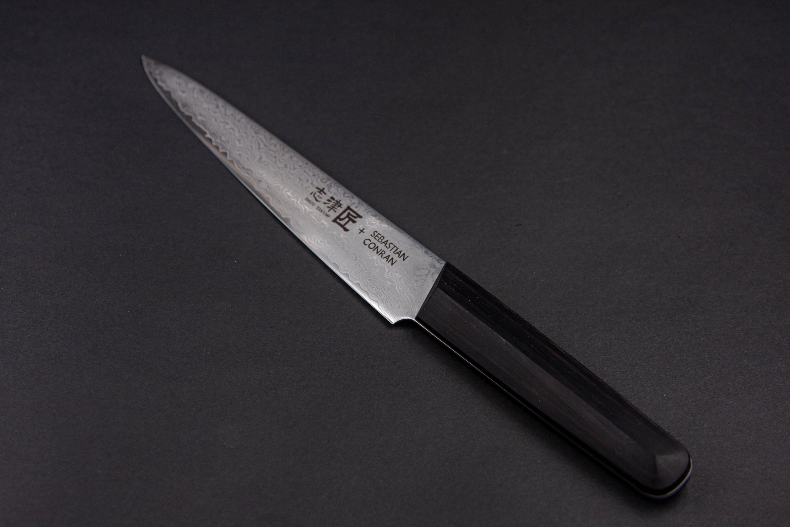 Das SC - Carving Knife zeichnet sich durch eine scharfe, gemusterte Stahlklinge und einen dunklen Holzgriff aus, der vor einem schwarzen Hintergrund im japanisch inspirierten Stil präsentiert wird.