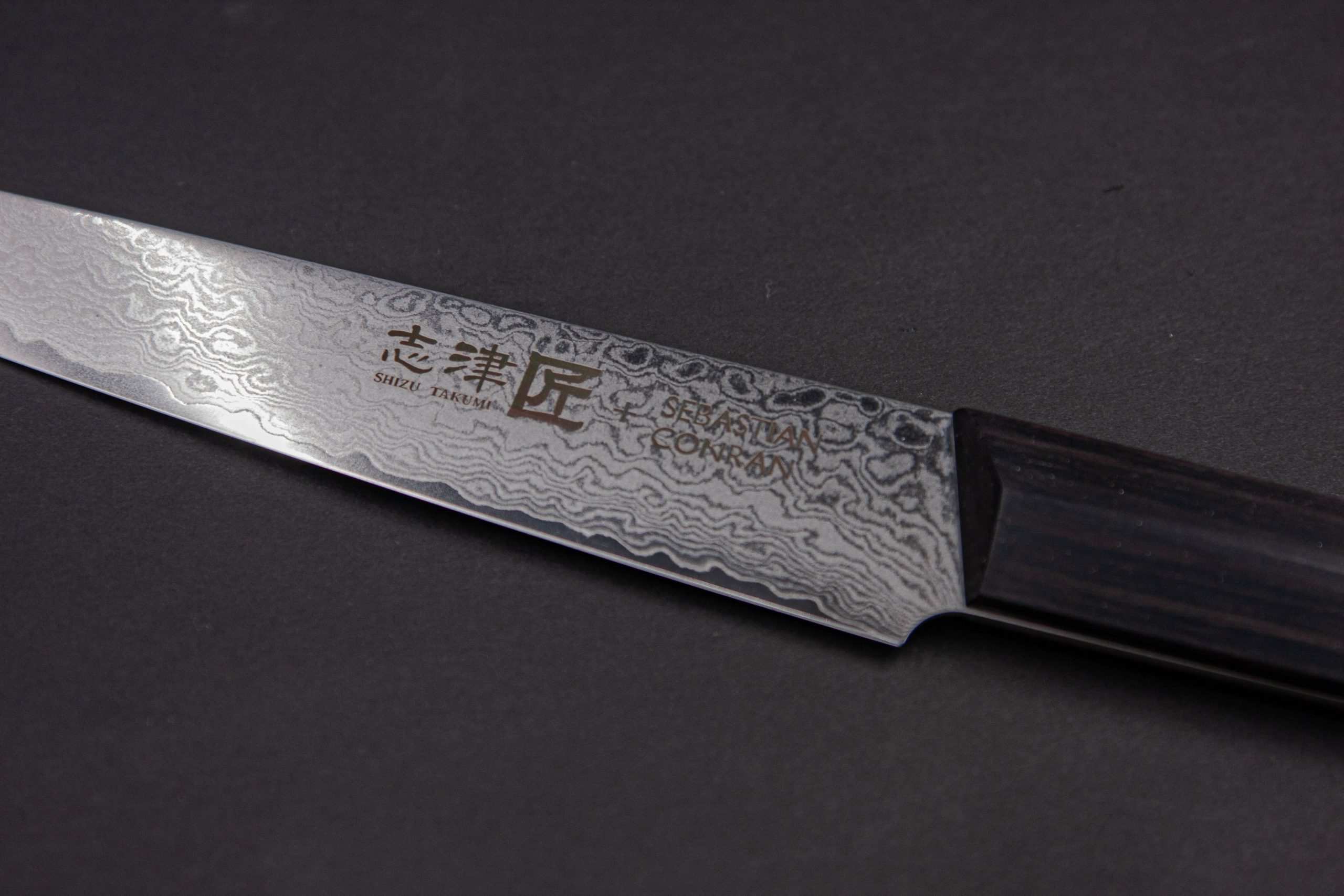 Eine Nahaufnahme des SC - Carving Knife zeigt den dunklen Holzgriff und die damaszierte Klinge mit eingravierten japanischen Schriftzeichen und englischem Text vor dunklem Hintergrund.