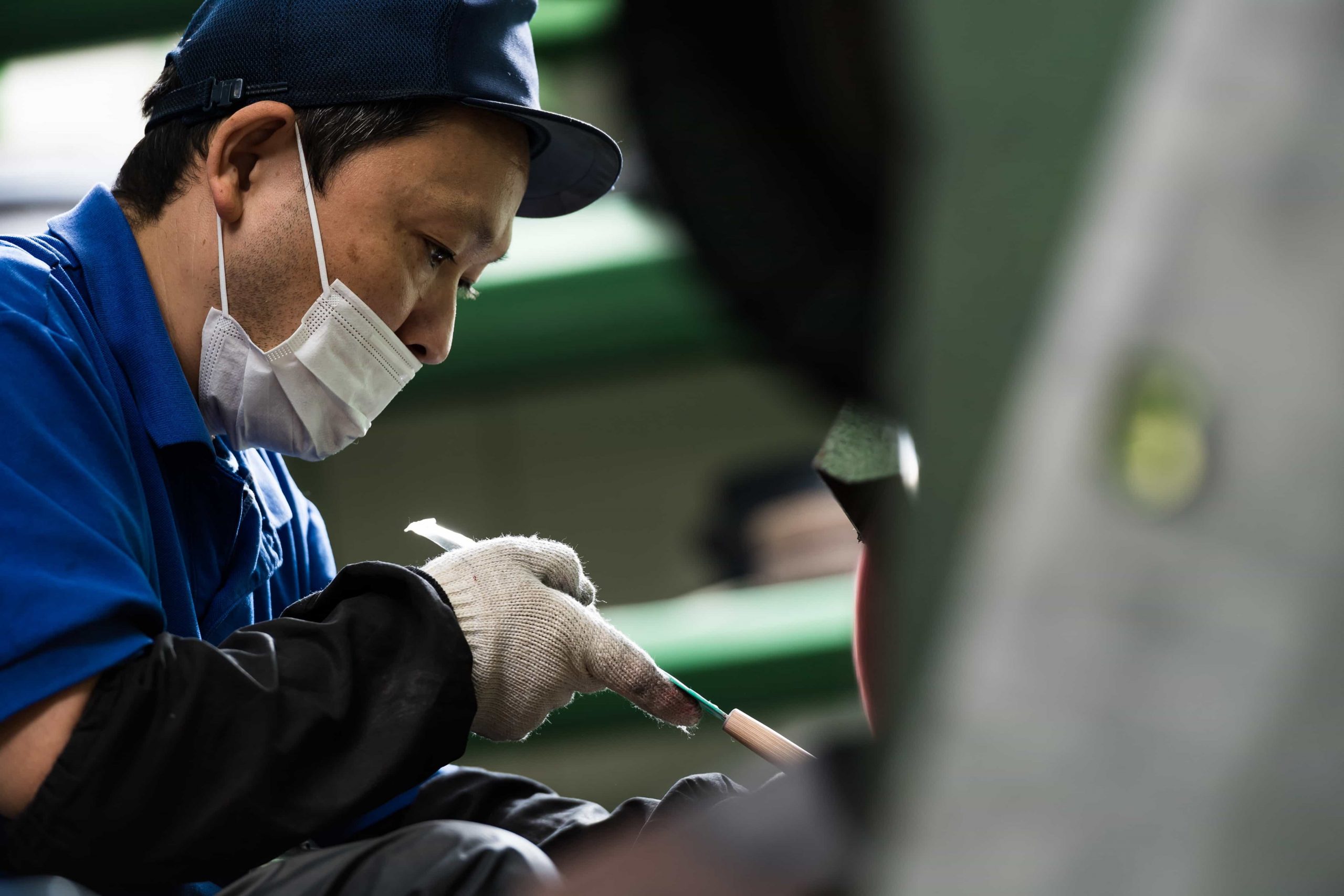 Ein Mann, der ein blaues Hemd, eine Mütze, einen Mundschutz und Handschuhe trägt, arbeitet sorgfältig mit seinen Händen und konzentriert sich auf eine Aufgabe in einer Industrie- oder Werkstattumgebung.
