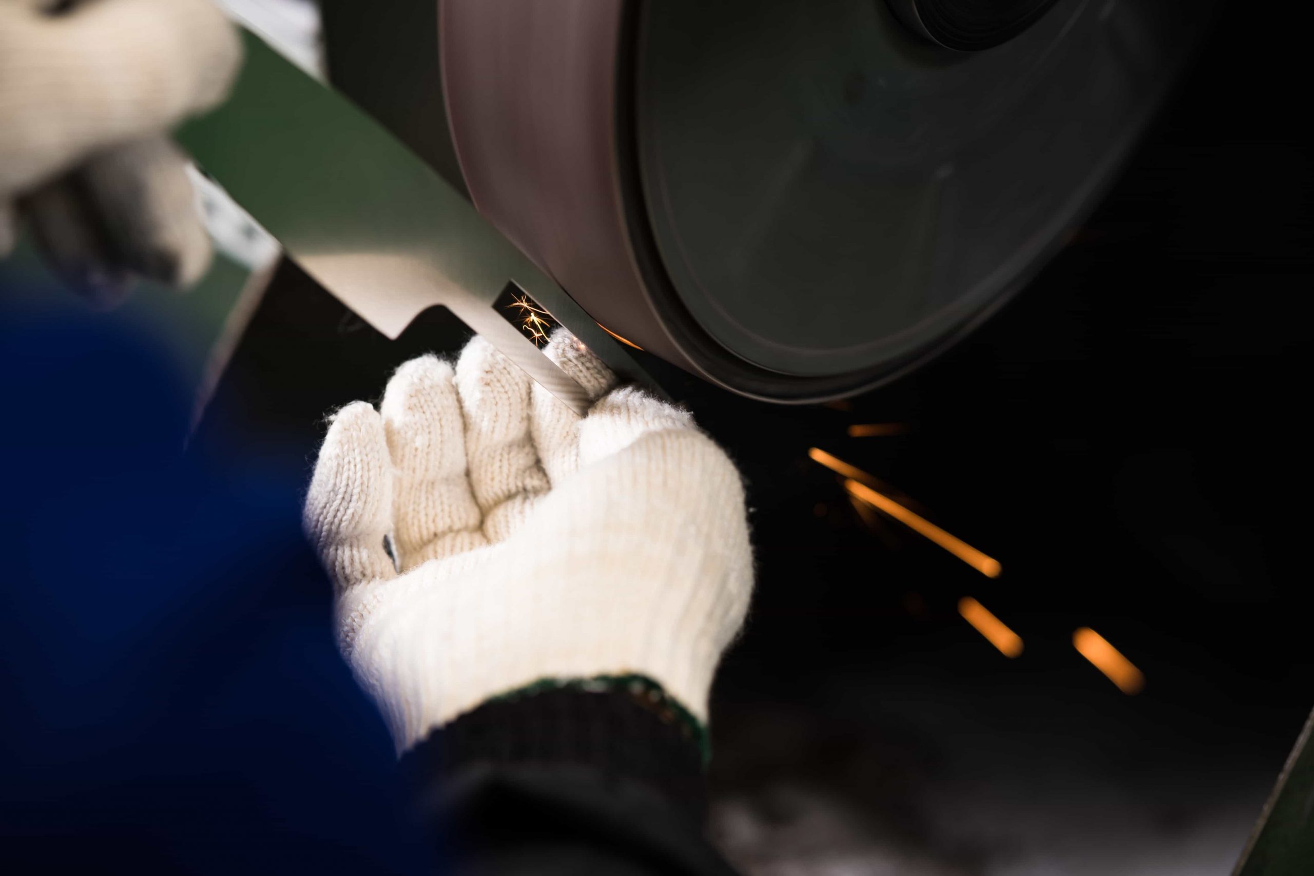 Eine Person mit weißen Handschuhen schärft eine Messerklinge auf einer Schleifscheibe, wobei Funken von der Kante fliegen.
