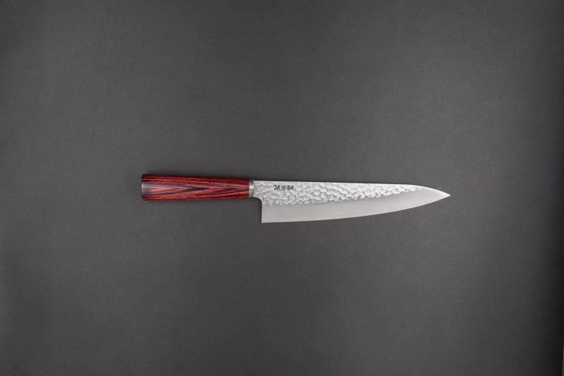 Ein japanisches Kochmesser mit gehämmerter Stahlklinge und einem dunklen Holzgriff ist horizontal auf einem schlichten dunklen Hintergrund platziert.
