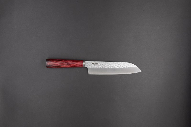 Ein Küchenmesser im japanischen Stil mit einer strukturierten silbernen Klinge und einem dunkelroten Holzgriff auf einem schlichten dunkelgrauen Hintergrund.