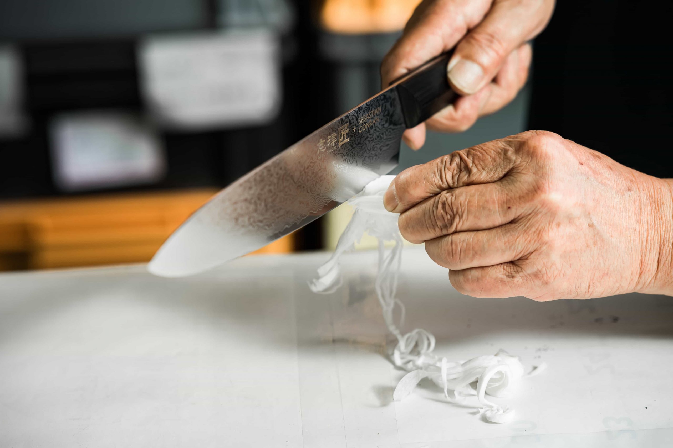 Eine Person verwendet ein scharfes Küchenmesser, um ein weißes Gemüse, möglicherweise eine Zwiebel, auf einem weißen Schneidebrett in feine Scheiben zu schneiden. Unter dem Messer sind dünne Streifen des Gemüses zu sehen.