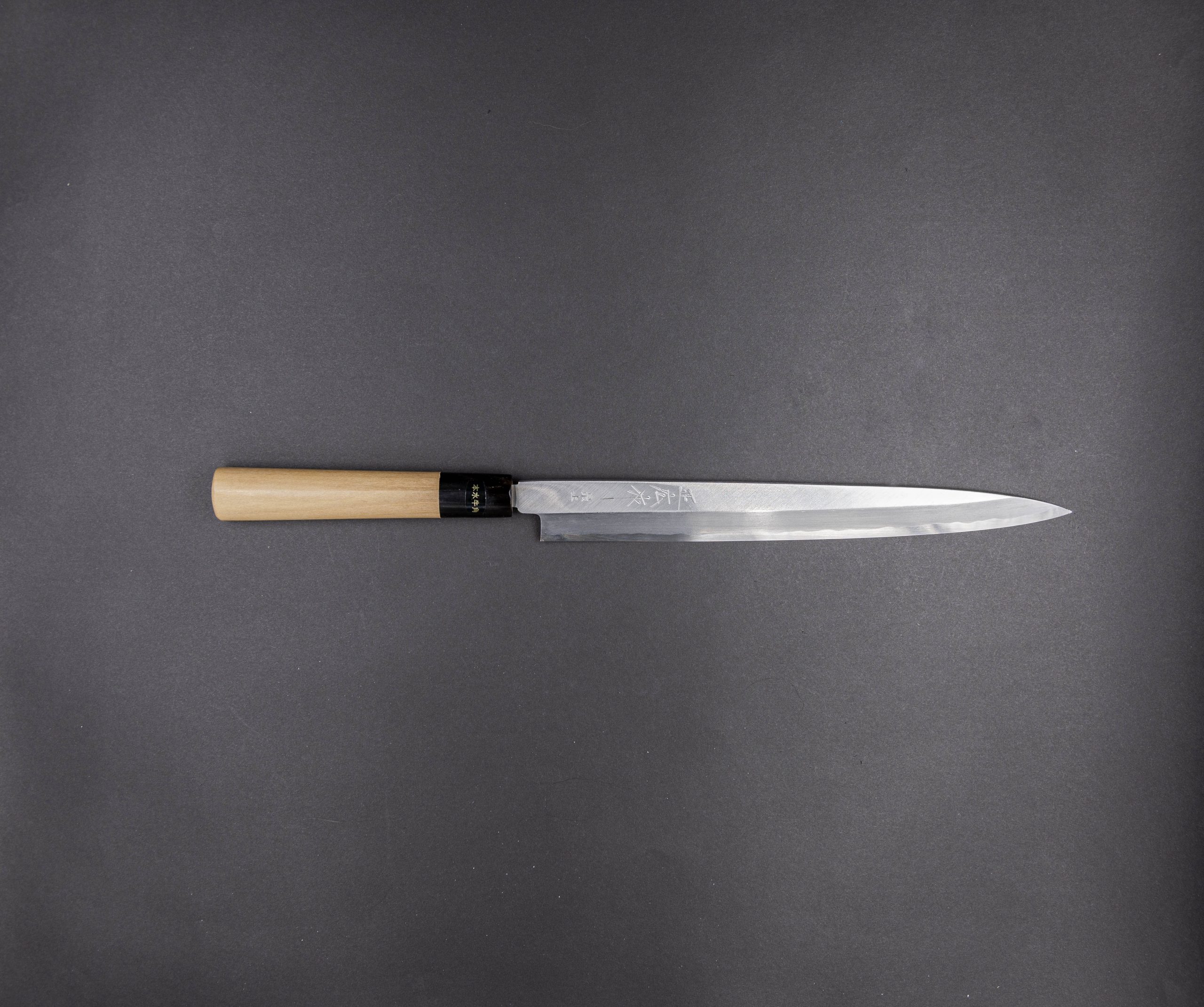 Ein einzelnes japanisches Kochmesser mit einem Holzgriff und einer langen, schlanken Klinge ist horizontal auf einem schlichten dunkelgrauen Hintergrund platziert.
