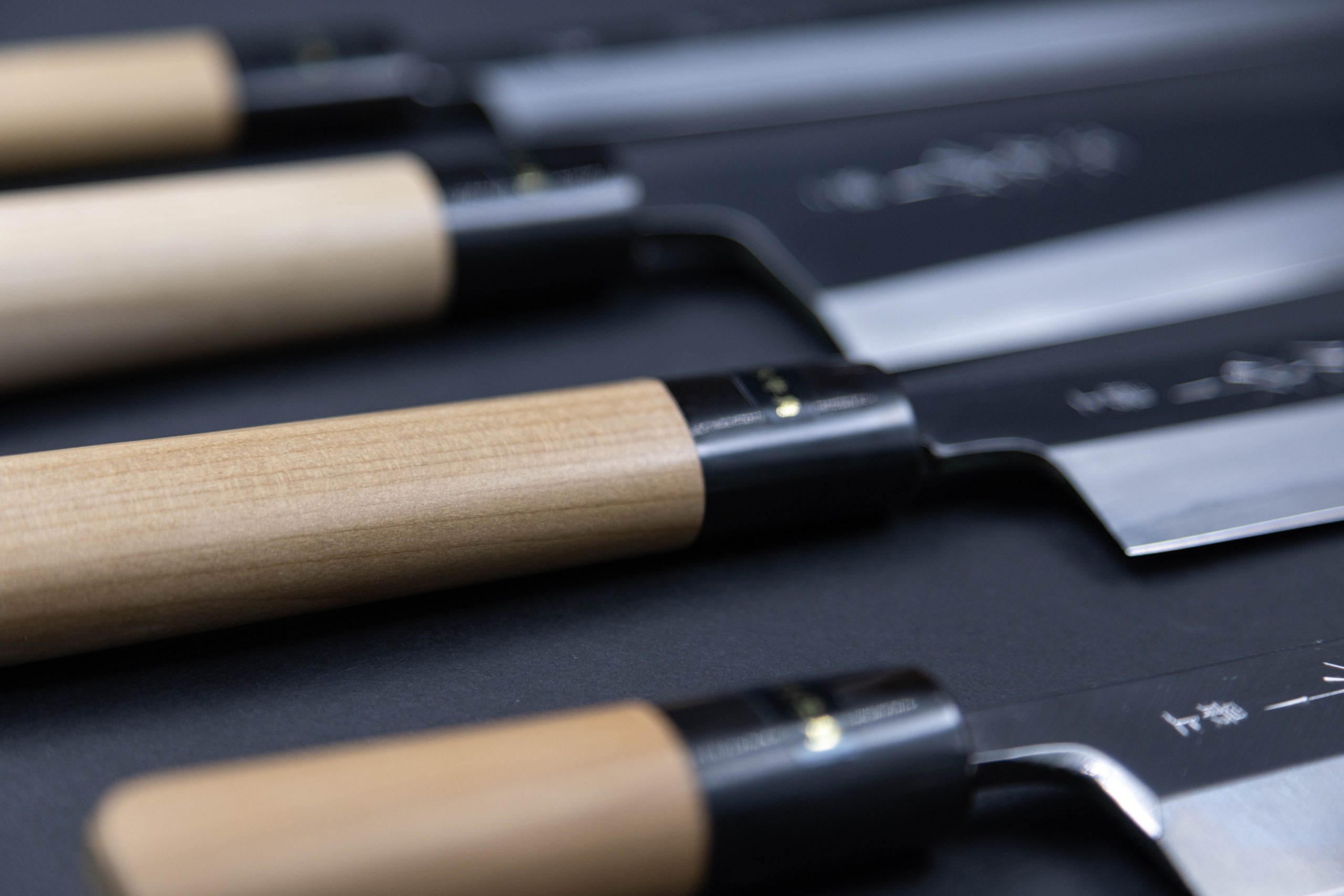 Eine Nahaufnahme mehrerer japanischer Küchenmesser mit hellen Holzgriffen und glänzenden Metallklingen, die parallel auf einer dunklen Fläche angeordnet sind.
