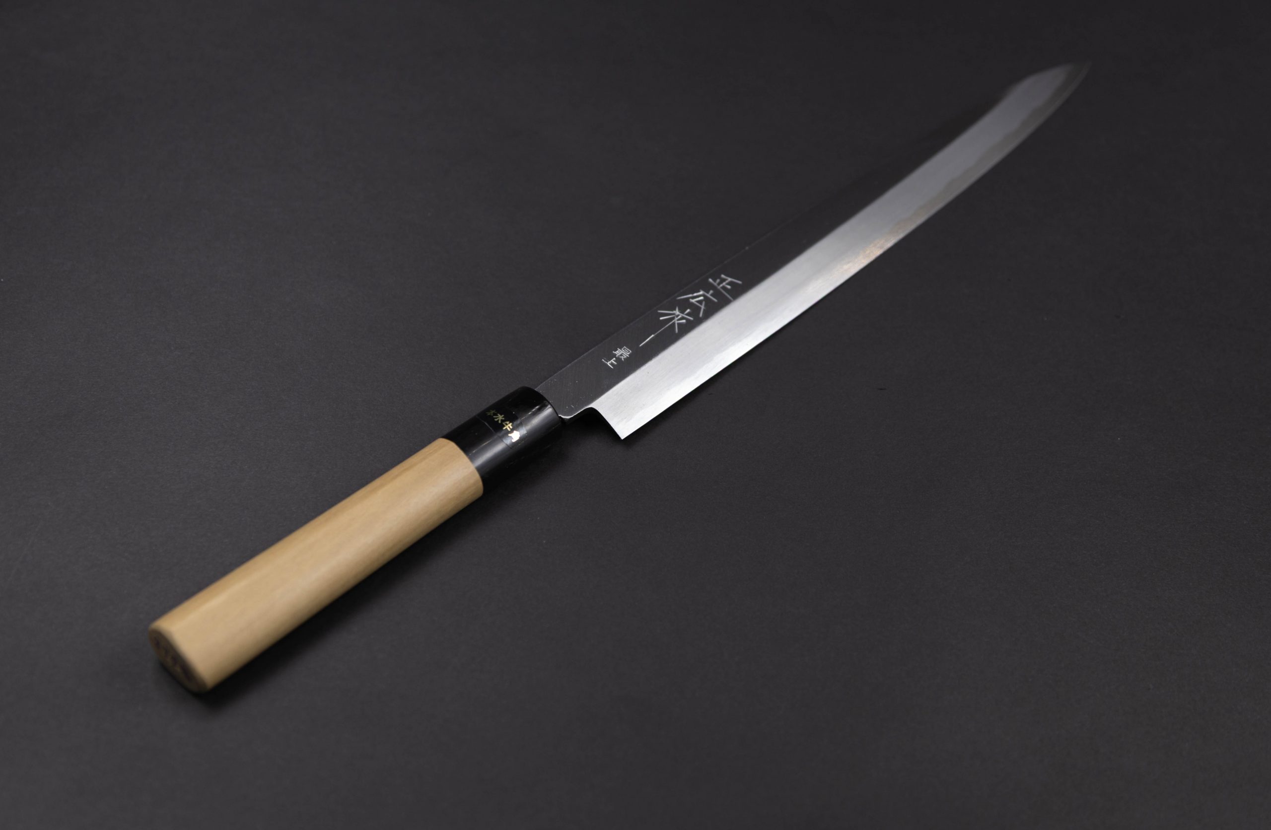 Ein japanisches Küchenmesser mit einem hellen Holzgriff und einer langen, schlanken Stahlklinge, die auf einer dunklen, glatten Oberfläche ausgestellt ist.