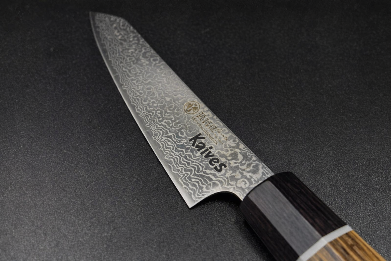 Nahaufnahme eines gemusterten Kochmessers mit dunklem Holzgriff, das auf einer schwarzen Fläche liegt. Auf der Klinge sind dekorative, wellenförmige Muster und das Wort "Knives" aufgedruckt.