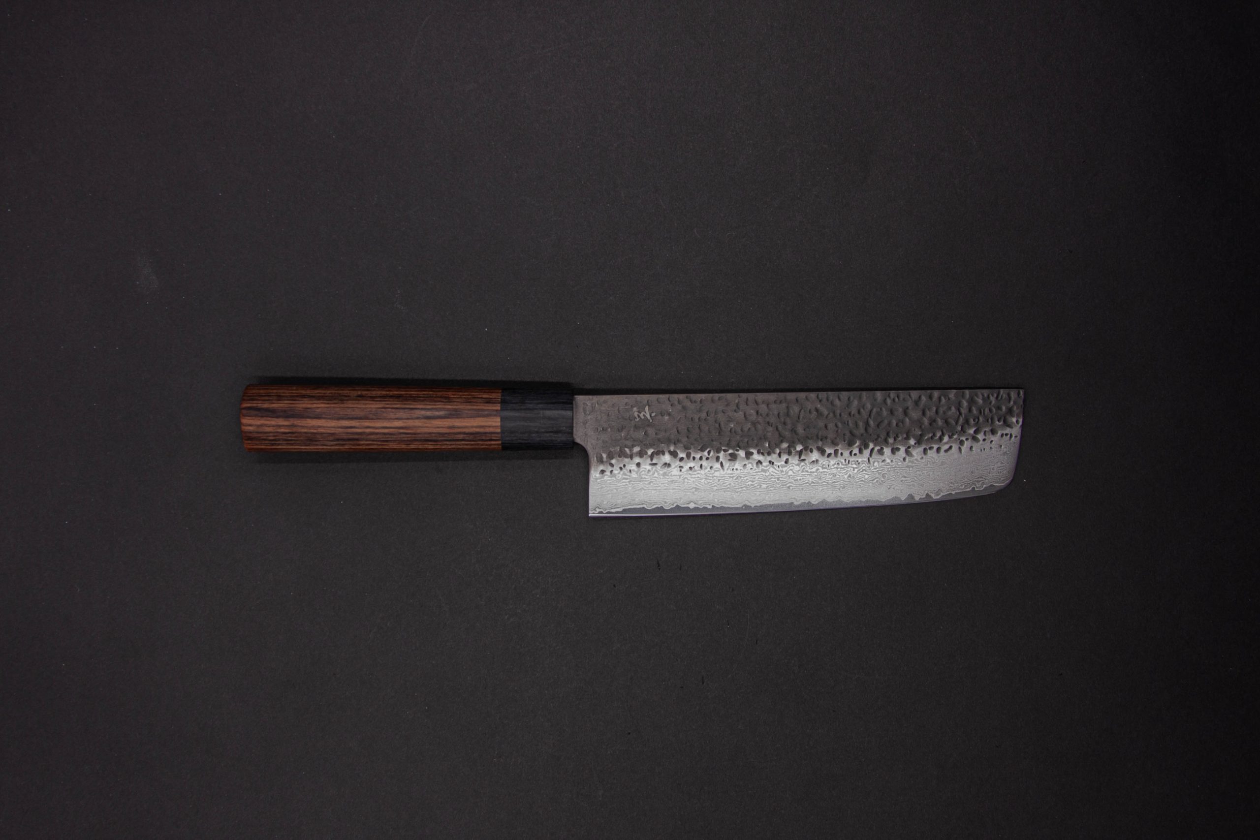 Das Gen - Nakiri 150mm, ein Küchenmesser im japanischen Stil mit dunklem Holzgriff und gehämmerter, strukturierter Klinge, wird auf einem schlichten, dunklen Hintergrund präsentiert.