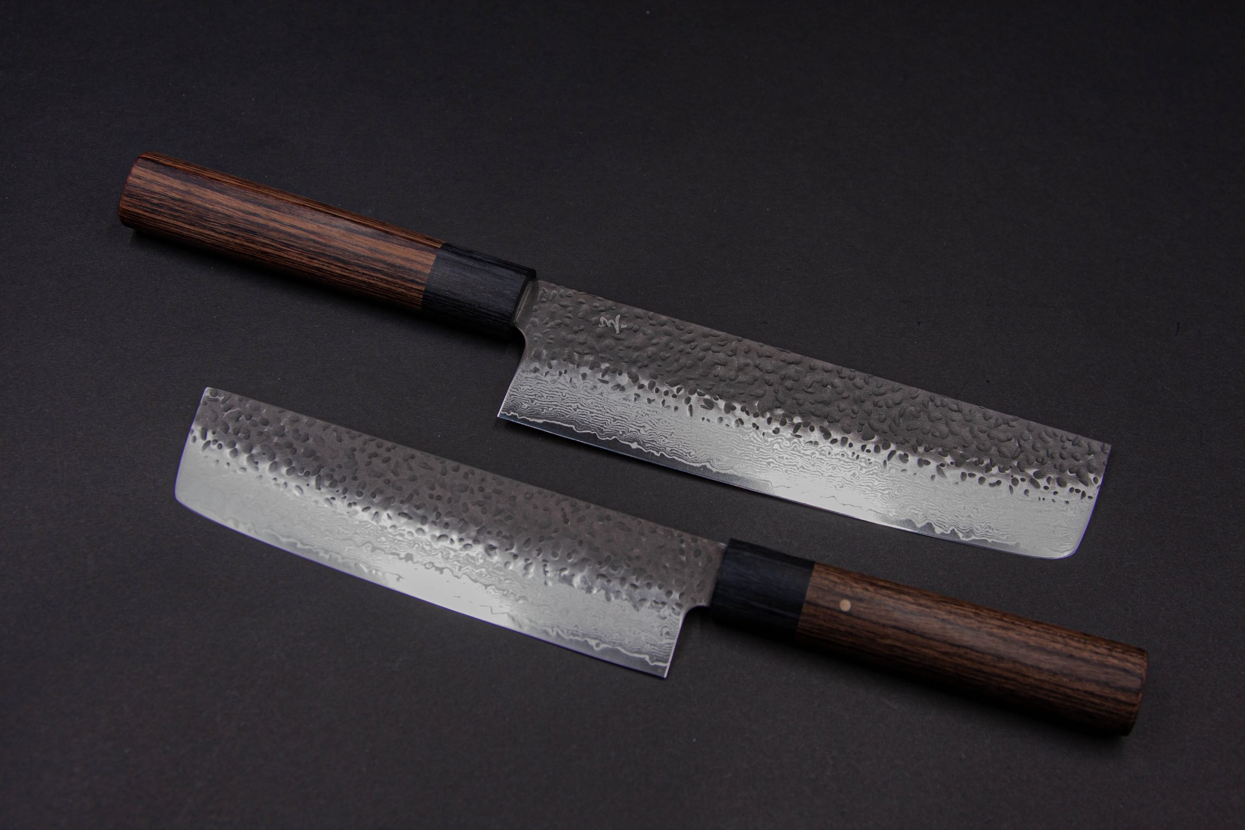 Das japanische Küchenmesser Gen - Nakiri 150 mm hat eine gehämmerte, strukturierte Stahlklinge und einen dunklen Holzgriff, der elegant auf einer dunklen Oberfläche angeordnet ist.