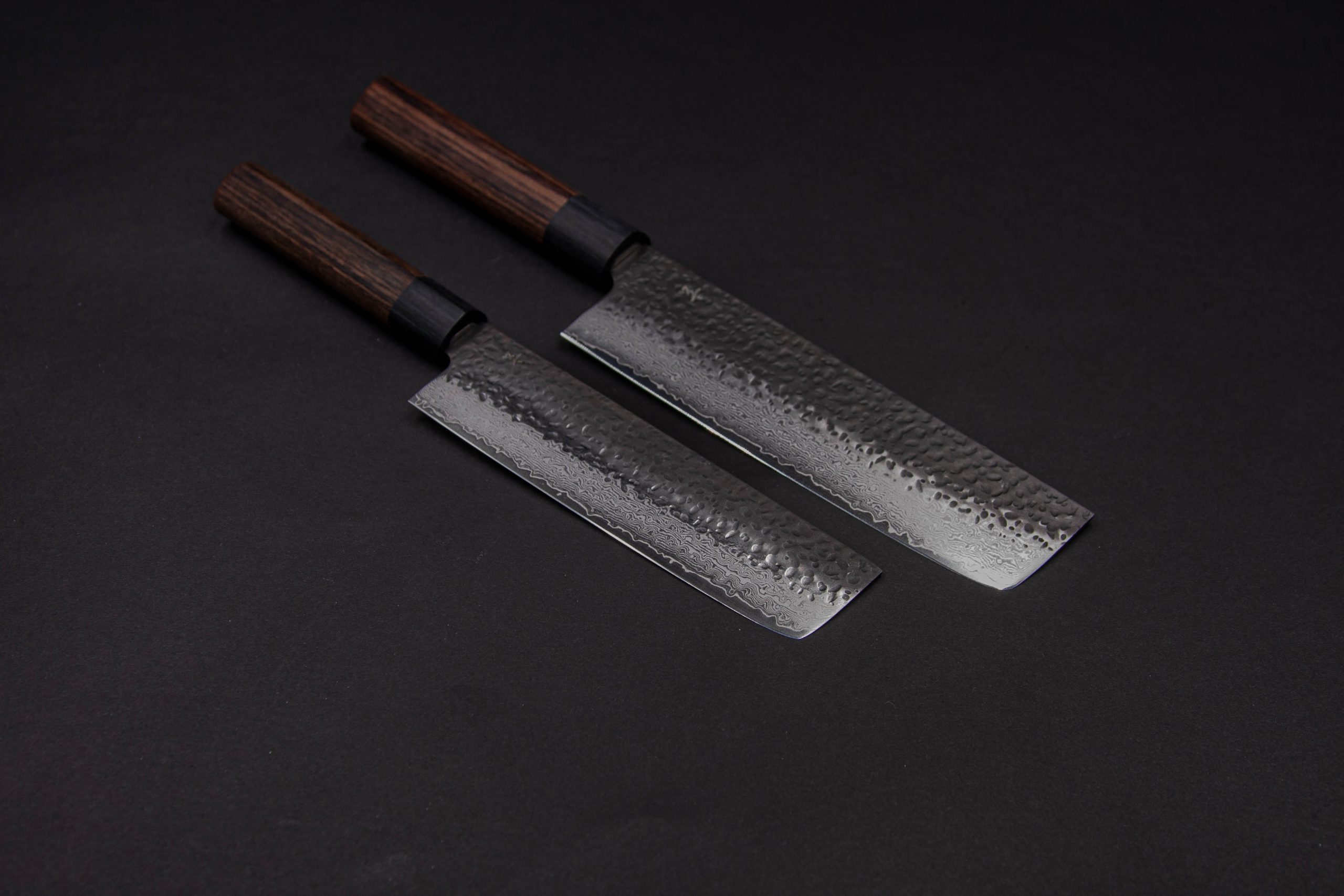 Zwei Küchenmesser im japanischen Stil, darunter das Gen-Nakiri 150 mm mit Holzgriff und gehämmerter Stahlklinge, liegen nebeneinander auf einer dunklen Fläche.