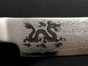 Nahaufnahme einer Messerklinge mit einem gewellten Damaszenerstahlmuster und einem eingravierten schwarzen Drachenmuster mit einem E. daneben. Der Hintergrund ist eine strukturierte schwarze Oberfläche.