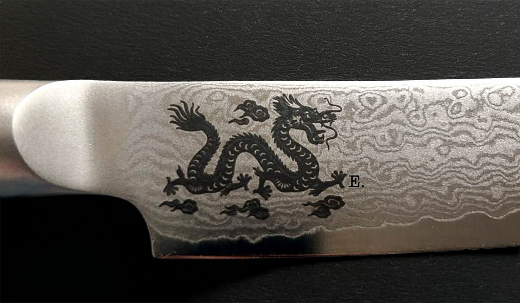 Nahaufnahme einer Messerklinge mit einem gewellten Damaszenerstahlmuster und einem eingravierten schwarzen Drachenmuster mit einem E. daneben. Der Hintergrund ist eine strukturierte schwarze Oberfläche.